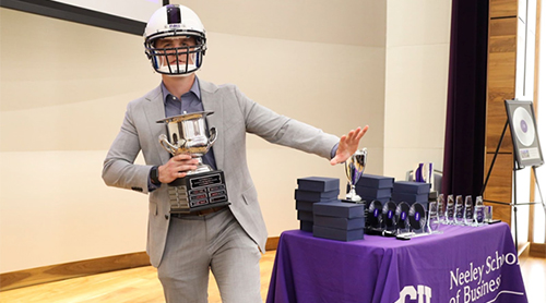 Student wearing a TCU football helmet holding a trophy by a table full of trophies.