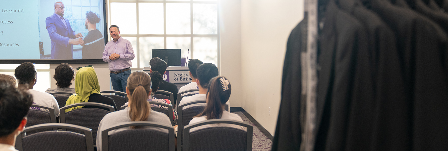 Section Image: Les Garrett addresses a group of students in the Neeley Banquet Room with a rack of suit coats in the back 